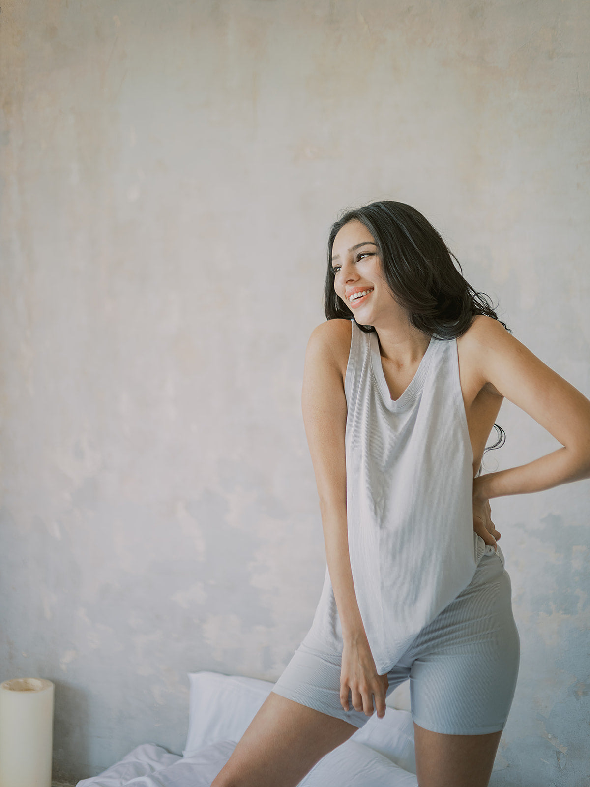 Woman in light gray tank top and shorts sitting on a bed with a neutral background